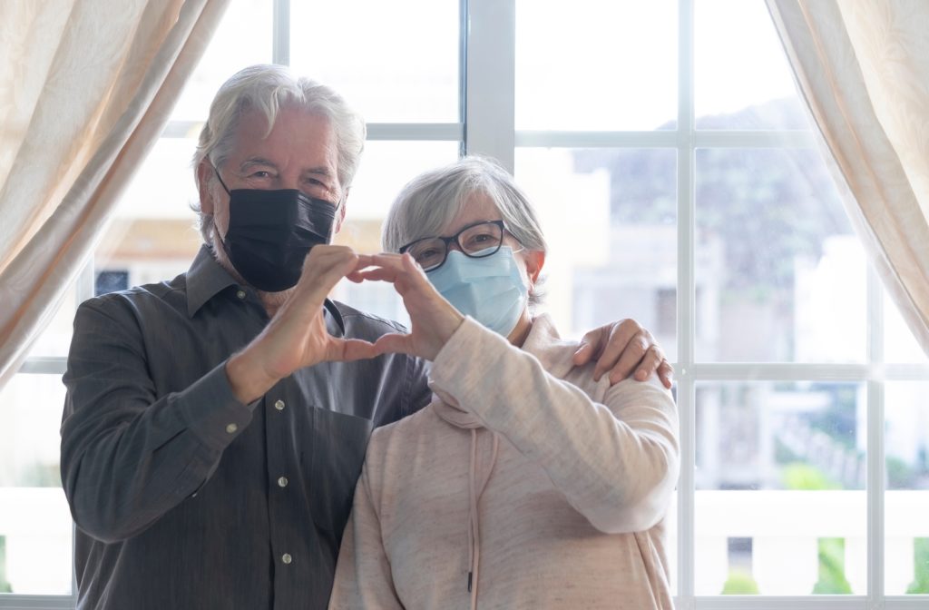 A senior couple standing beside each other with the husband's arm around his wife while they create a heart shape by joining each other's hands together. They are both wearing surgical mask's in front of a large window