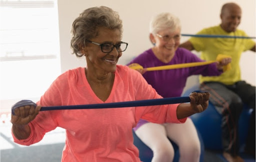 Senior woman in a group exercise class sitting on an exercise ball stretching exercise bands across her chest. Behind her are a male and female senior smiling and doing the same.
