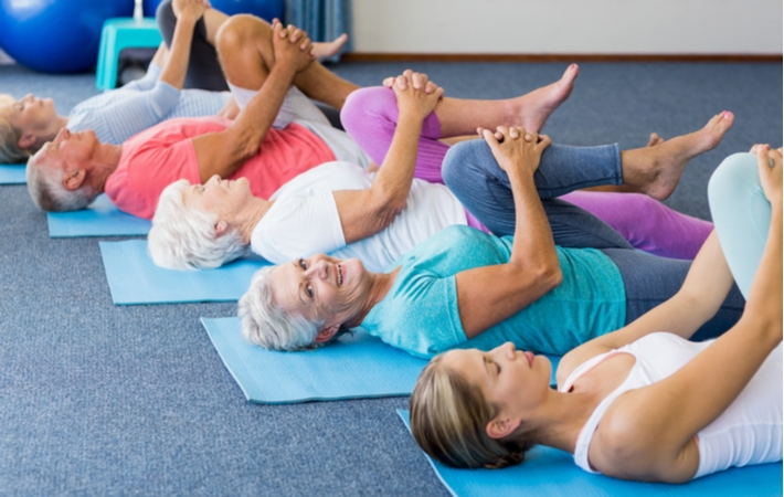 Группа пожилых людей, лежащих на ковриках для йоги, прижимающих колени к груди, следуя примеру женщины-инструктора, лежащей рядом с ними.