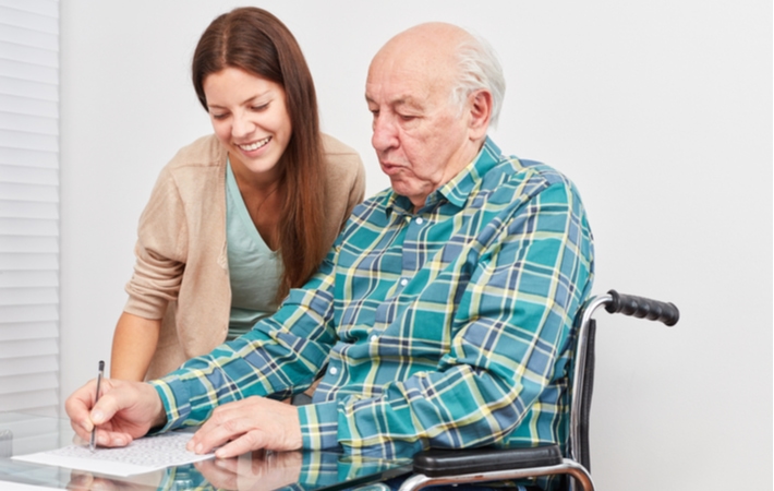 A senior man in a wheelchair sitting at a table doing memory care activities with a caregiver helping him over his shoulder