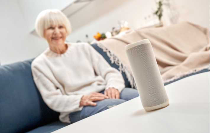 A senior woman sitting on a sofa smiling, looking towards a smart speaker