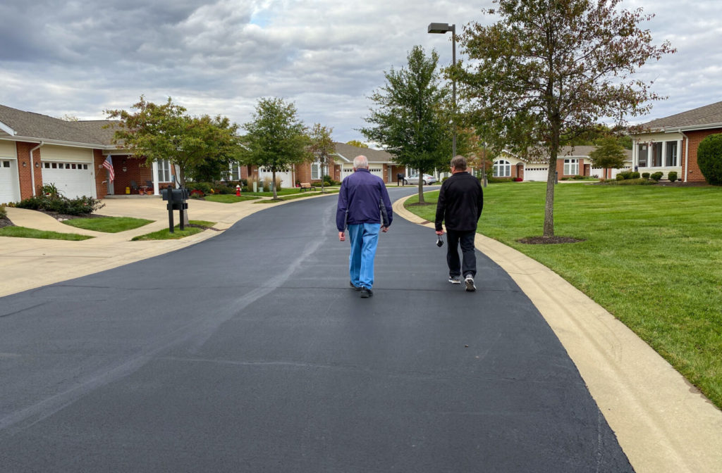 "A son walking with his father on the road of a senior living community with many trees and houses."