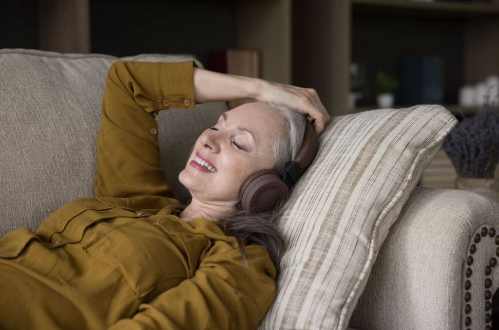 A smiling older adult lying on a couch with their eyes closed and listening to music with wireless headphones.