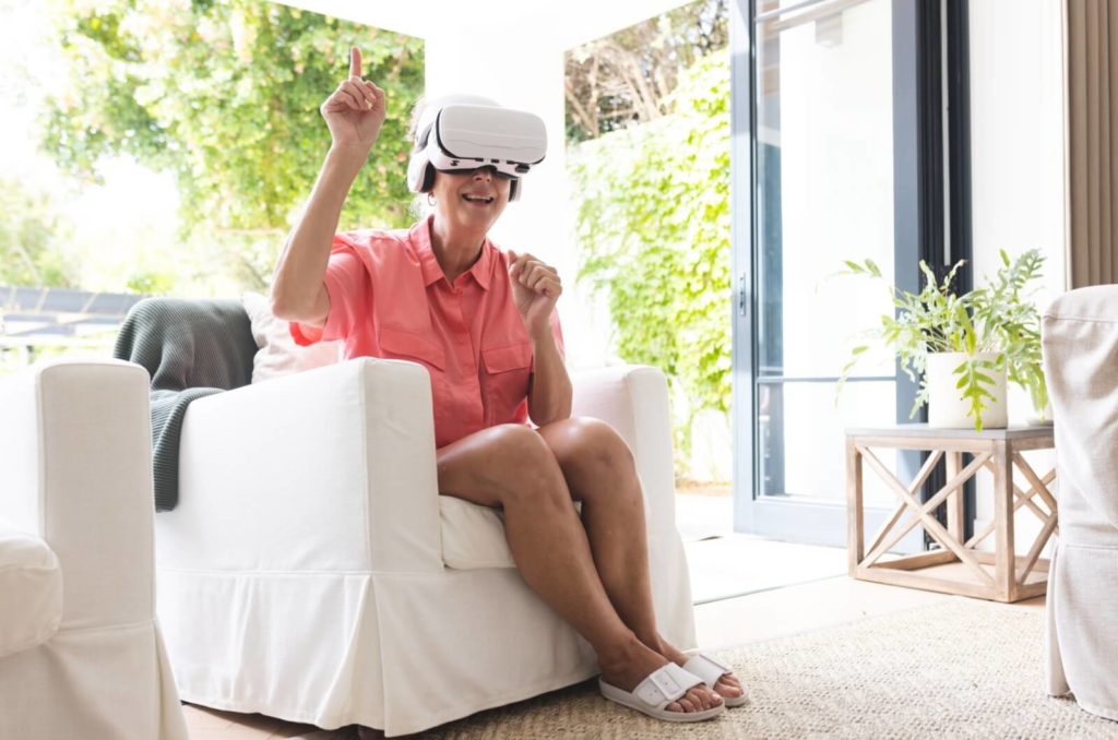 A smiling older adult sitting in a white armchair in a bright and airy room wearing a Virtual Reality headset.